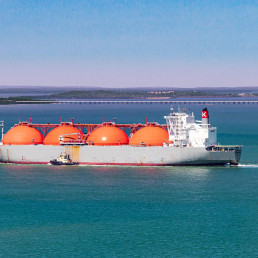 Ein Tanker mit vier runden orangenen Gastanks auf azurblauem Wasser nahe einer Küste.